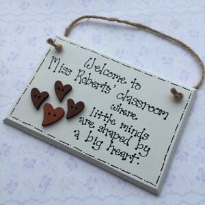 May include: White wooden sign with brown heart-shaped buttons and the text "Welcome to Miss Roberts' classroom where little minds are shaped by a big heart."