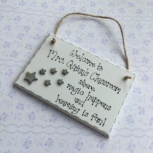 May include: White wooden sign with the text "Welcome to Mrs. Quinn's Classroom where magic happens and learning is fun!" with silver glitter stars.