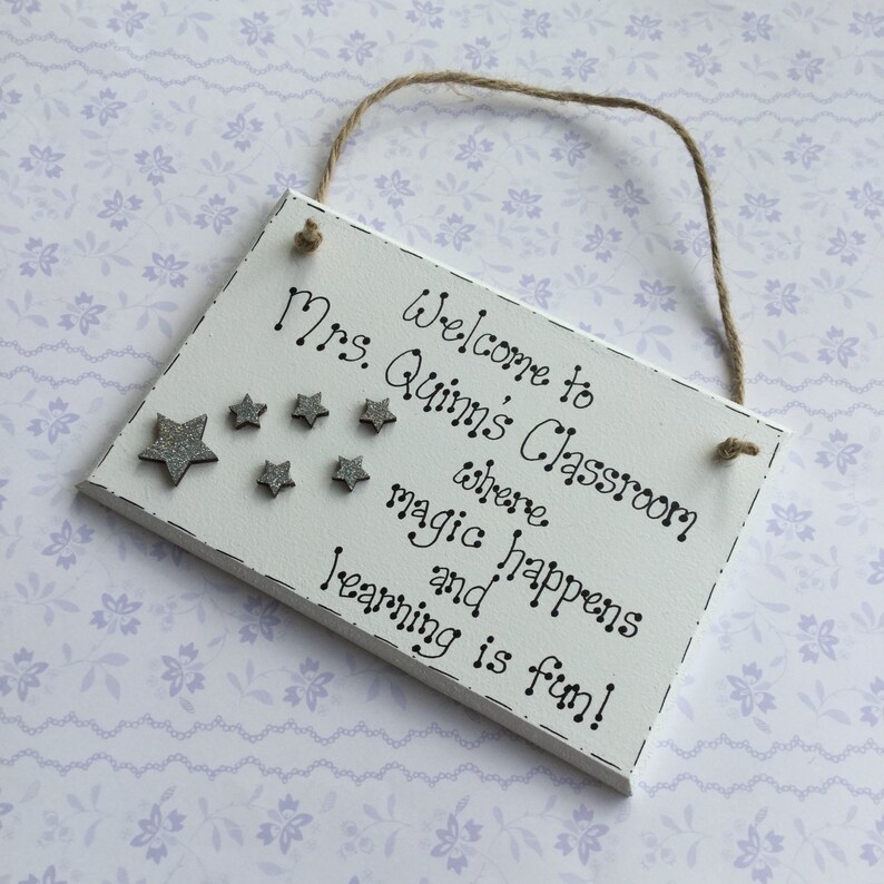 May include: White wooden sign with the text "Welcome to Mrs. Quinn's Classroom where magic happens and learning is fun!" with silver glitter stars.