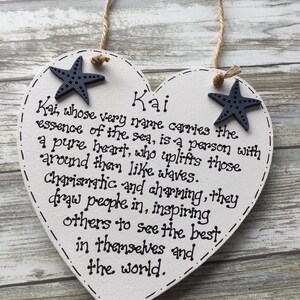 Puede incluir: Cartel de madera blanco en forma de coraz&oacute;n con el nombre "Kai" y una descripci&oacute;n escrita en tinta negra. Dos estrellas de mar azul marino adornan las esquinas superiores. Una cuerda de yute est&aacute; unida para colgar.