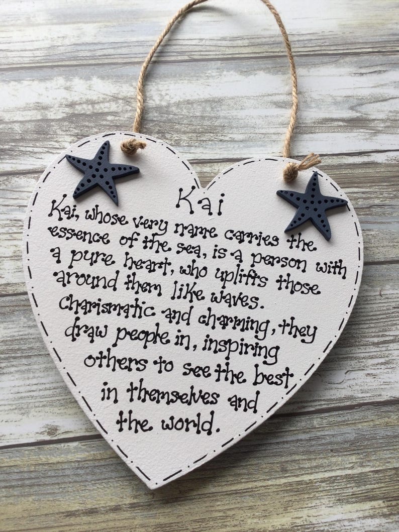Puede incluir: Cartel de madera blanco en forma de coraz&oacute;n con el nombre "Kai" y una descripci&oacute;n escrita en tinta negra. Dos estrellas de mar azul marino adornan las esquinas superiores. Una cuerda de yute est&aacute; unida para colgar.