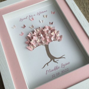 May include: A framed artwork featuring a paper tree with a canopy of pink butterflies. The artwork includes the text "Loved for a Lifetime", the name "Maddie Laine", and the date "12.08.2022". The frame is white with a pink mat.