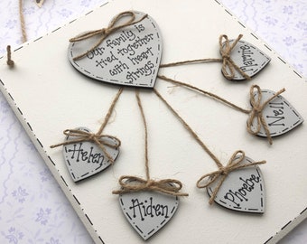 May include: White wooden plaque with the text "Our family is tied together with heart strings" and five smaller white wooden hearts hanging from it with twine. The hearts are labelled "Helen", "Aiden", "Phoebe", "Nell", and "Jamie".