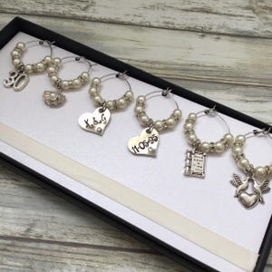 May include: A set of six silver-toned wine glass charms featuring white faux pearls and clear rhinestones. The charms include dangling pendants shaped like the number 30, a seashell, a heart with K&G, a heart with 11-05-95, a book, and two birds.