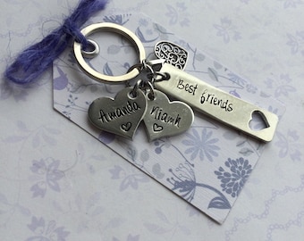 May include: A silver-toned metal keychain featuring a circular ring with a purple fiber tie. Attached are two heart-shaped charms engraved with the names Amanda and Niamh, a rectangular bar charm stamped with the text Best friends, and a small filigree heart.