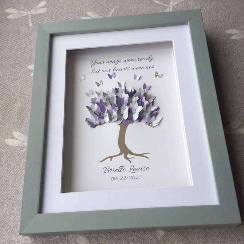 May include: A framed artwork featuring a tree with a butterfly-filled canopy in shades of purple, silver, and white. The tree trunk is brown, and the frame is gray. Text reads, "Your wings were ready, but our hearts were not." Below the tree is the name "Brielle Louise" and the date "09/22/2023."