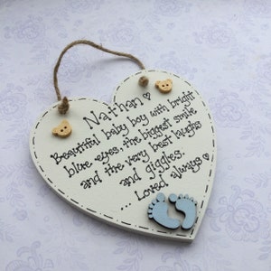 Puede incluir: Placa de madera en forma de coraz&oacute;n crema con el nombre "Nathan" y un mensaje sobre un beb&eacute;. Presenta letras negras, botones de oso de madera y huellas de pies azules. Se adjunta una cuerda de yute para colgar.