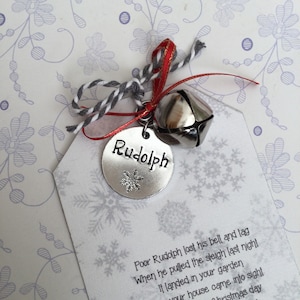 May include: A silver tag with the name "Rudolph" and a snowflake design, attached to a silver bell with a red ribbon and a white and gray twine bow. The tag has a poem about Rudolph losing his bell and tag.
