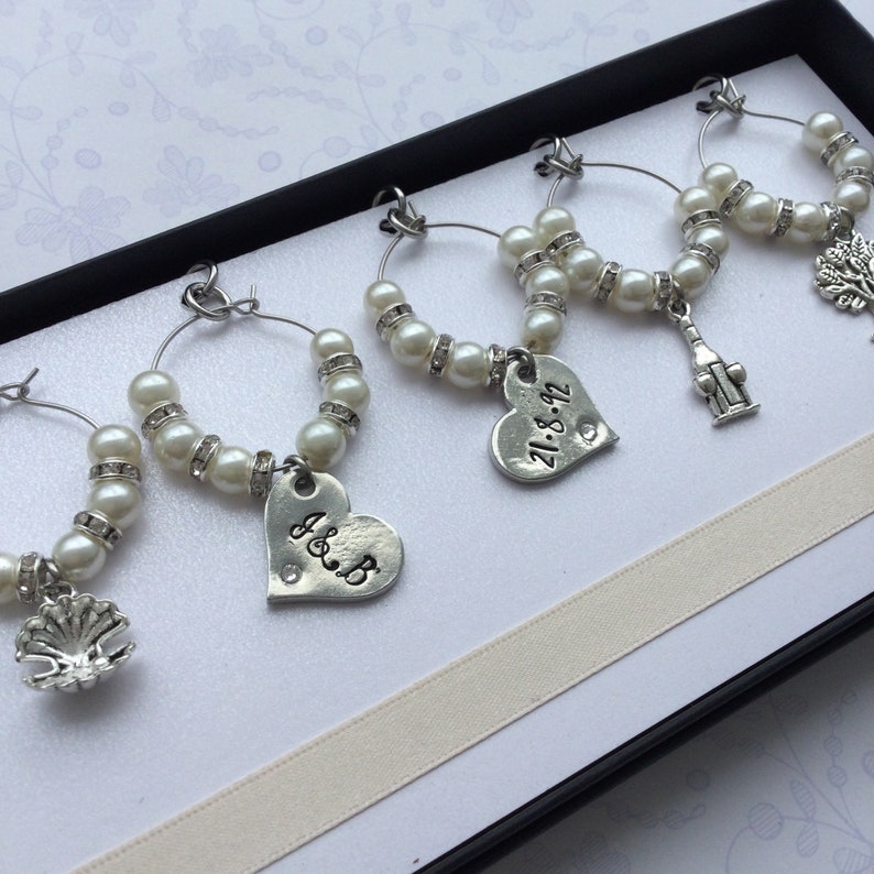 May include: A set of four silver-toned wine glass charms featuring white faux pearls and clear rhinestones. Each charm includes a unique metal pendant: a seashell, a heart stamped with J&B, a heart stamped with 21.8.92, and a tree.