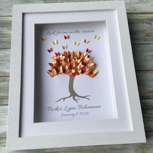 May include: A framed paper art piece featuring a tree with a brown trunk and a colorful canopy of orange, yellow, and red leaves. Butterflies flutter around the tree. The text "Look for me in the sunrise" is above the tree, and the name "Parker Lynn Bohannan" and date "January 9 2026" are below.