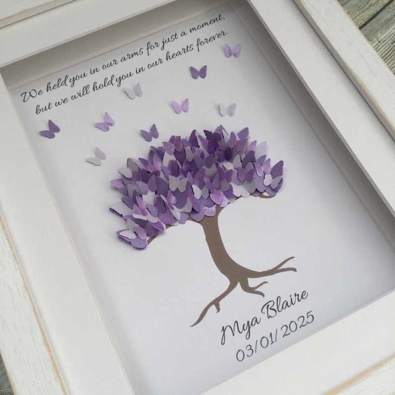 May include: A framed art piece featuring a tree with a purple butterfly-shaped canopy. The tree trunk and roots are brown. The artwork includes the text "We held you in our arms for just a moment, but we will hold you in our hearts forever." and the name "Mya Blaire" with the date "03/01/2025".