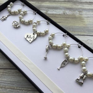 May include: A set of six silver-toned wine glass charms arranged in a black box. Each circular hoop features white faux pearls and clear rhinestones. The charms include a shell, a heart engraved with A&G, a wine glass, a flute, and a cheers sign.
