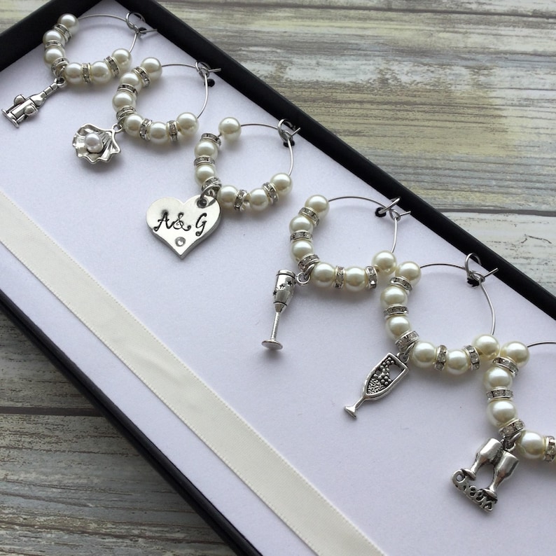 May include: A set of six silver-toned wine glass charms arranged in a black box. Each circular hoop features white faux pearls and clear rhinestones. The charms include a shell, a heart engraved with A&G, a wine glass, a flute, and a cheers sign.