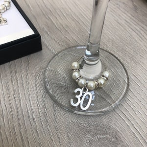 May include: A wine glass charm featuring a silver-toned number 30 pendant attached to a circular wire ring. The ring is decorated with alternating white faux pearls and small clear crystal-encrusted silver spacers, designed to wrap around a glass stem.