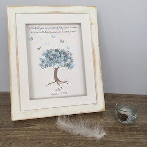 May include: A framed art print with a white distressed frame. The artwork features a tree with blue and gray leaves and butterflies. The text reads, "We held you in our arms for just a moment, but we will hold you in our hearts forever." Includes initials and a date.