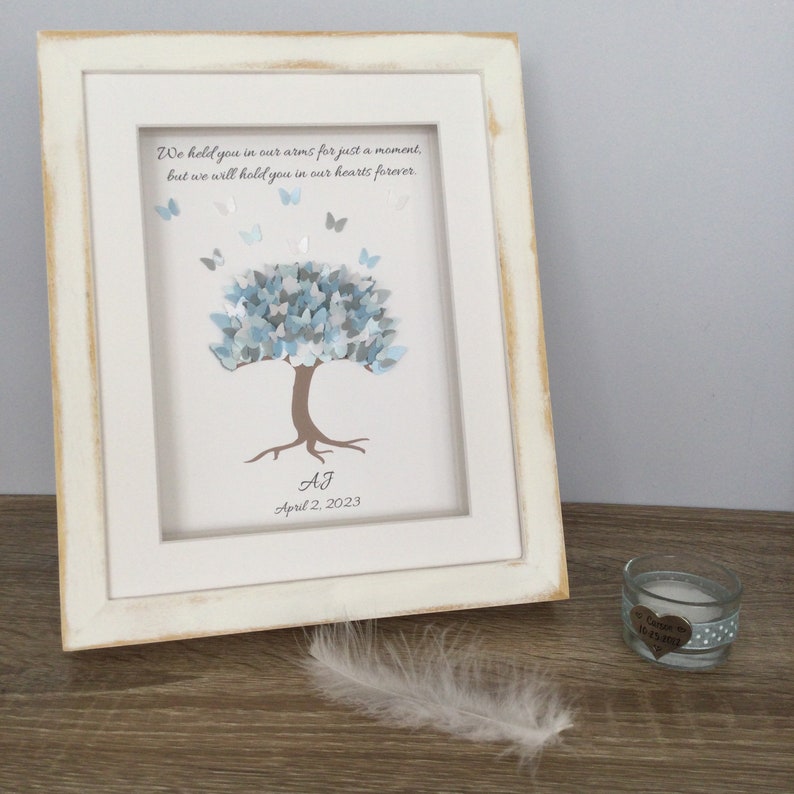 May include: A framed art print with a white distressed frame. The artwork features a tree with blue and gray leaves and butterflies. The text reads, "We held you in our arms for just a moment, but we will hold you in our hearts forever." Includes initials and a date.
