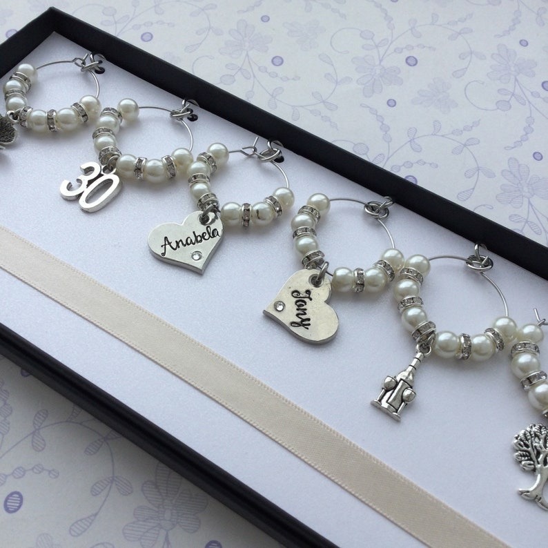 May include: A set of six silver-toned wine glass charms arranged in a black box. Each circular charm features white faux pearls, small clear rhinestones, and unique silver pendants including the number 30, heart shapes with names Anabela and Tony, a tower, and a tree.