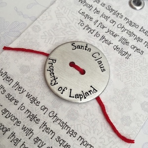 May include: A silver button with the text "Santa Claus Property of Lapland" and a red thread attached to it. The button is on a white card with the text "This is Santa's magic button which he lost on Christmas night. Leave it for your little ones to find to their delight."