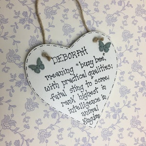Puede incluir: Un letrero de madera blanco en forma de coraz&oacute;n con el nombre "DEBORAH" y su significado escrito en negro. Tiene adornos de mariposas y un colgador de cuerda. El fondo es un estampado floral.