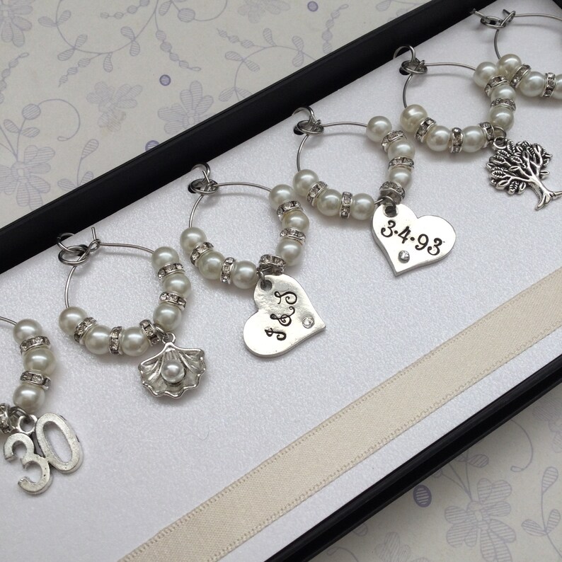 May include: A set of five silver-toned wine glass charms featuring white faux pearls and clear rhinestones. Each charm includes a unique metal pendant: the number 30, a seashell, a heart with J&S, a heart with 3.4.93, and a tree of life charm.