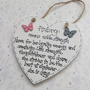 Puede incluir: Placa de madera en forma de coraz&oacute;n con el nombre "Aubry" y su significado, junto con una descripci&oacute;n de las caracter&iacute;sticas del nombre. Cuenta con adornos de mariposas rosas y azules y una cuerda de yute para colgar.
