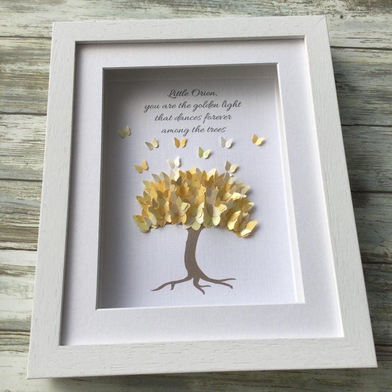 May include: A framed artwork featuring a paper tree with golden butterfly leaves and a brown trunk. The artwork includes the text "Little Orion, you are the golden light that dances forever among the trees." The frame is white.