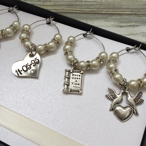 May include: A set of silver-toned wine glass charms featuring white faux pearls and clear rhinestones. The charms include dangling metal pendants: a heart engraved with 11-05-95, a book charm with the text Once Upon A Time, and two doves above a heart.