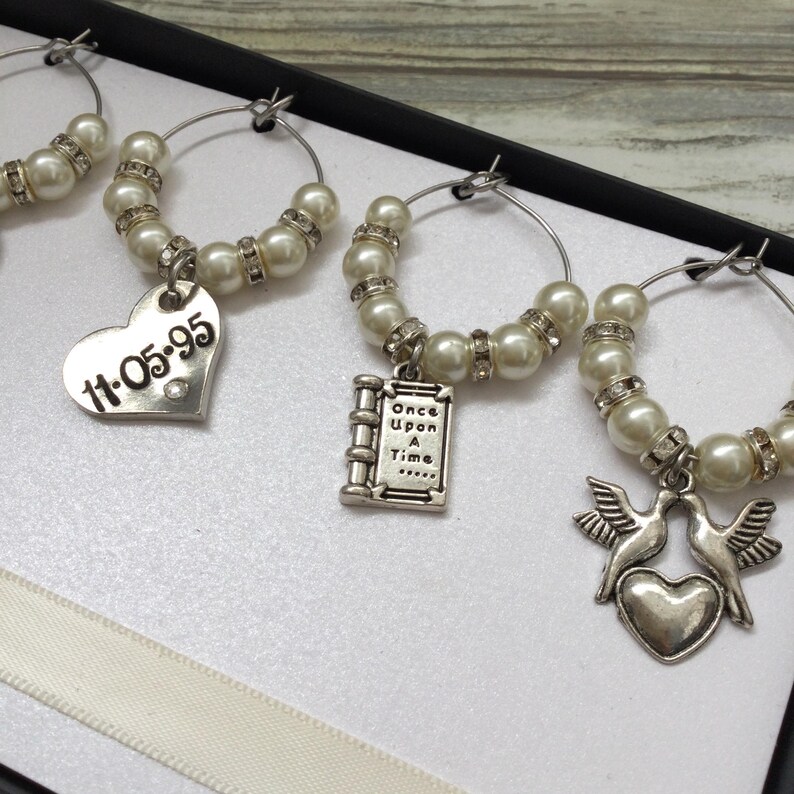 May include: A set of silver-toned wine glass charms featuring white faux pearls and clear rhinestones. The charms include dangling metal pendants: a heart engraved with 11-05-95, a book charm with the text Once Upon A Time, and two doves above a heart.