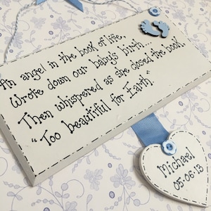 May include: A white wooden plaque with a blue ribbon and a heart-shaped tag. The plaque has a handwritten message that reads "An angel in the book of life, Wrote down our baby's birth... Then whispered as she closed the book, "Too beautiful for Earth." The heart-shaped tag has the name "Michael" and the date "05-06-13" written on it.