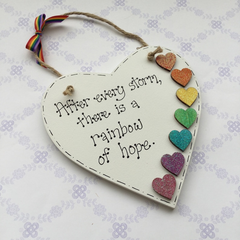 May include: White wooden heart-shaped sign with the text "After every storm, there is a rainbow of hope."  The sign is decorated with a rainbow of glitter hearts.