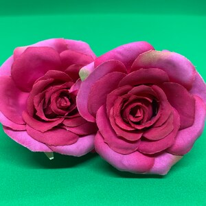 Peut inclure: Deux roses en tissu rose avec un design de p&eacute;tales superpos&eacute;s. Les roses sont attach&eacute;es &agrave; un bandeau noir.