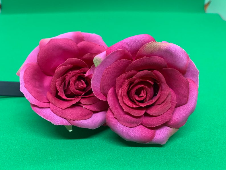 Peut inclure: Deux roses en tissu rose avec un design de p&eacute;tales superpos&eacute;s. Les roses sont attach&eacute;es &agrave; un bandeau noir.