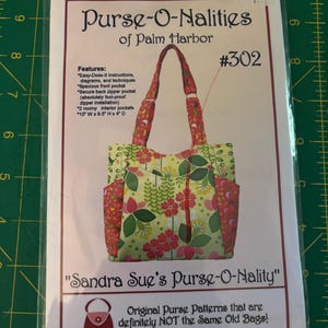 May include: A sewing pattern for a tote bag, titled "Purse-O-Nalities of Palm Harbor" by Sandra Sue. The pattern features a floral design in red, green, and pink. The bag measures 10" W x 9.5" H x 4" D.