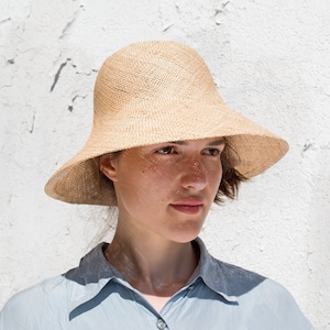 Könnte beinhalten: Eine Person trägt einen breitkrempigen, hellbraunen Strohhut mit leicht abgerundeter Krone. Der Hut hat eine gewebte Textur und eine breite, weiche Krempe.