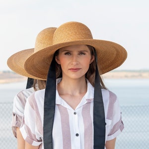 以下が含まれることがあります： 黒のリボンタイとピンクと白のストライプのボタンダウンシャツを着た女性。帽子はユニークなダブルドーム型です。