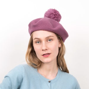 Può includere: Un cardigan blu chiaro con chiusura a bottoni. La modella indossa un berretto rosa con un pompon soffice.