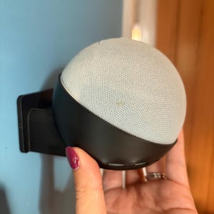 May include: A spherical smart speaker with a light grey fabric mesh cover, securely mounted in a black plastic wall bracket. The device features a rounded design with a visible horizontal seam where the fabric meets the black base housing.