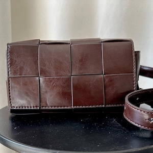 May include: A dark brown rectangular crossbody bag featuring a woven grid pattern with wide, smooth panels. The bag includes a matching adjustable strap with a metal buckle and visible stitching along the edges of the woven squares.