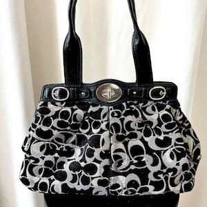 May include: A black and silver handbag featuring a repeating circular logo pattern across the fabric body. The bag includes black leather handles, a silver-toned metal turn-lock closure at the center, and decorative silver buckles on the top trim.