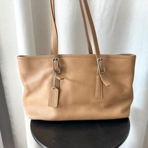 May include: A tan leather tote bag featuring two long shoulder straps with silver-toned metal buckles. A rectangular leather hang tag embossed with the word COACH is attached to one of the straps by a small silver-toned metal chain.