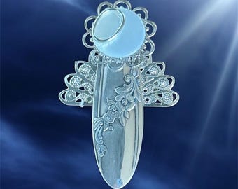 Puede incluir: Un objeto decorativo plateado en forma de ángel con un orbe azul claro en la parte superior. El ángel presenta intrincadas alas de filigrana y un diseño floral en el cuerpo. El fondo es un degradado de azul.
