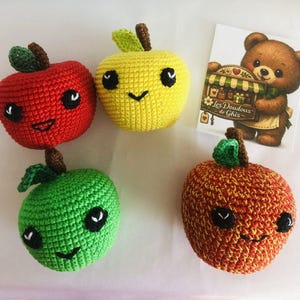 Op de afbeelding: Vier gehaakte speelgoed in de vorm van appels in rood, geel, groen en oranje. Elke appel heeft een lachend gezicht met zwarte ogen en een gebogen mond. Er is ook een kleine kaart met een berenillustratie en de tekst "Les Doudous de Ghis".