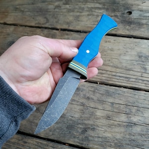 May include: A handcrafted knife with a textured, dark gray blade and a vibrant blue handle. The handle features a white dot and is accented with yellow and green stripes. The knife is held against a wooden background.