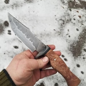 May include: A handcrafted hunting knife with a textured, silver blade and a wooden handle. The blade has a unique, wavy pattern. The handle is light brown with natural wood grain. A small, silver pin is visible on the handle.