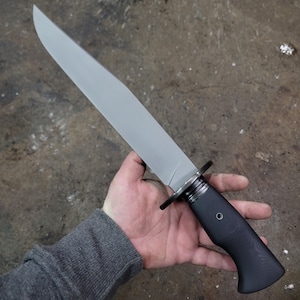 May include: A large, silver-bladed knife with a black handle. The blade is long and straight, with a sharp point. The handle is textured and curved for grip. A metal guard separates the blade and handle. The knife is held in a hand.