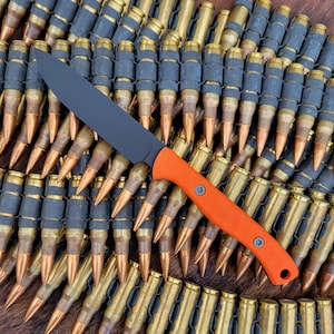 May include: A black and orange hunting knife with a black handle and a sharp blade. The knife is lying on top of a pile of ammunition.