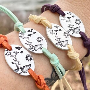 May include: Four bracelets with silver charms featuring a mountain scene with a sun, trees, and birds. The bracelets are made with different colored cords: orange, light green, cream, and purple.