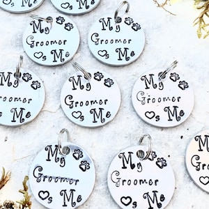 May include: A collection of silver-colored pet tags. Each tag is round and features the text "My Groomer Loves Me" in black script, along with paw prints and a heart symbol. A small metal ring is attached for easy attachment.