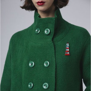 Puede incluir: Un abrigo de lana verde de doble botonadura con cuello alto y seis botones. Un pequeño amuleto de faro está prendido en la solapa.