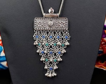 May include: A silver necklace with a rectangular pendant featuring intricate floral designs and blue and green gemstones. The pendant is attached to a delicate chain with a layered design.